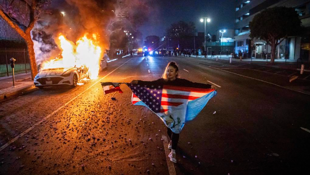 一名女子9日晚间拿着国籍在市中心示威,路过一辆燃烧中的汽车。 一名女子9日晚间拿着国籍在市中心示威,路过一辆燃烧中的汽车。